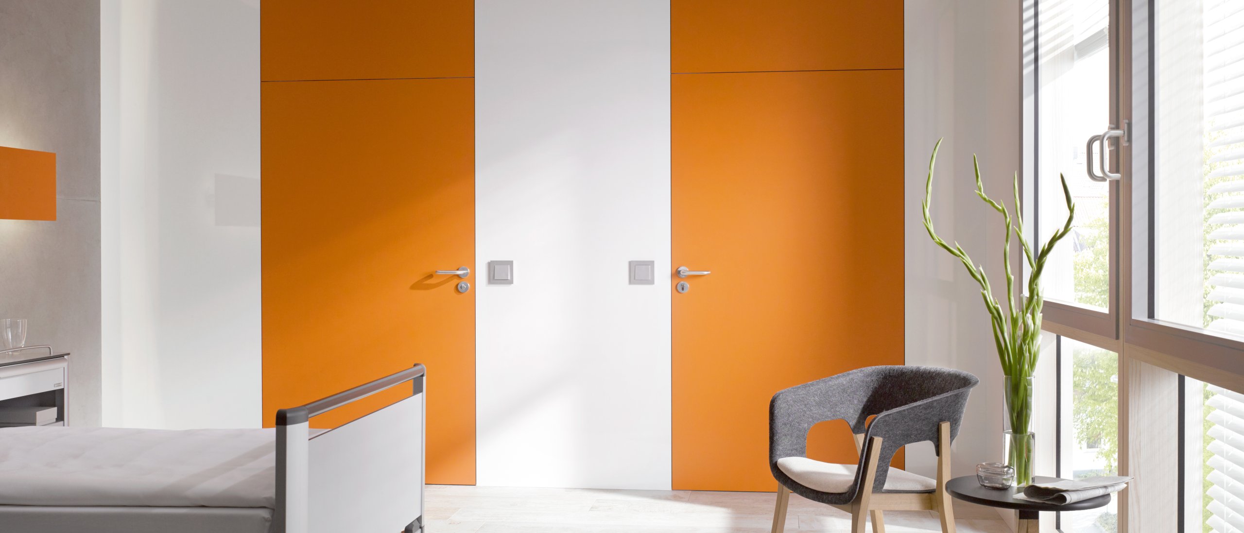 Weißes Krankenhauszimmer mit zwei orangenen Türen ausgestattet mit einem Türdrücker in der Farbe Grau aus Polyamid